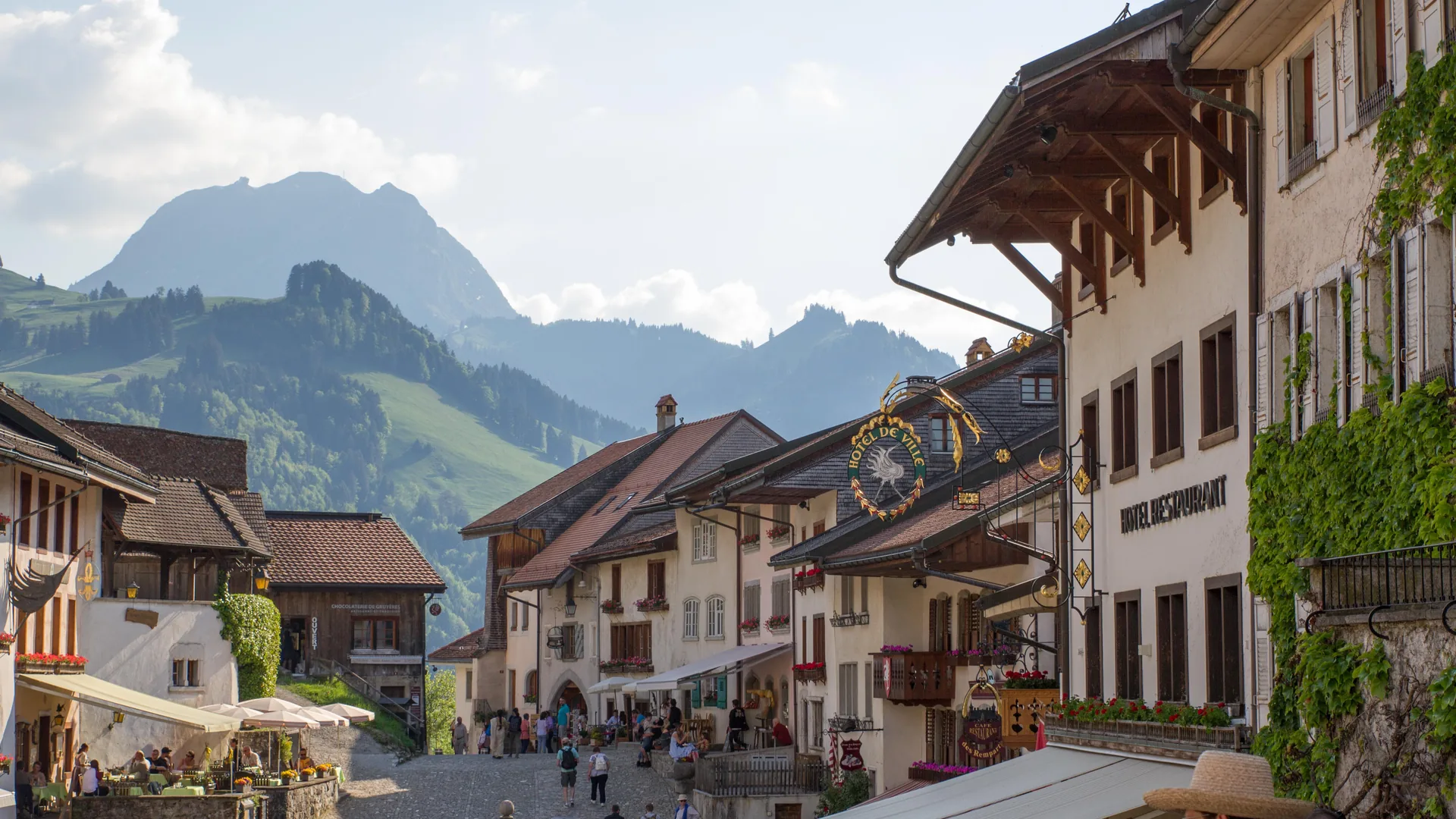 The cobbled main street of Gruyères with old houses, café terraces and a green mountain rising behind, from the Gruyères tour listing